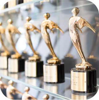 A row of shiny golden trophies with abstract human figures, displayed on glass shelves. The trophies have black bases with engraved plaques. The background is softly blurred, emphasizing the elegance of the awards.