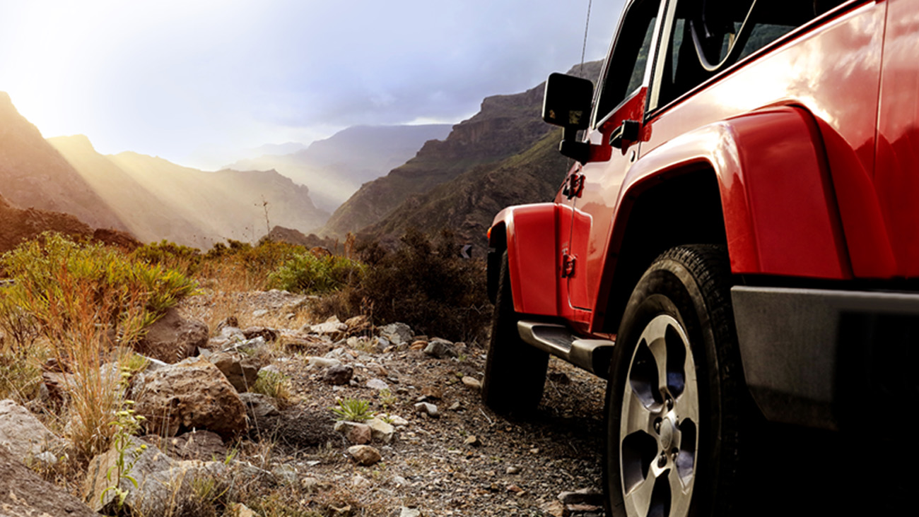 A red off-road vehicle is parked on a rugged, rocky trail surrounded by mountainous terrain. Sunlight streams through cloud cover, illuminating the landscape with a warm glow. Vegetation is sparse along the trail.