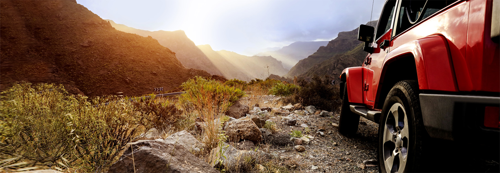 A red off-road vehicle driving on a rocky mountain trail, surrounded by rugged, sunlit terrain. Sun rays pierce through the cloudy sky, casting light on the mountainous landscape.