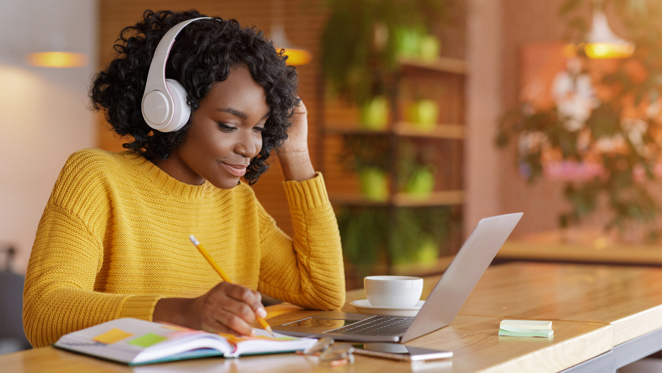 A woman wearing headphones is sitting at a wooden table with a laptop, cup, and notebook. Shes writing with a pencil, surrounded by plants and soft lighting. She appears focused, with a hand resting on her head.