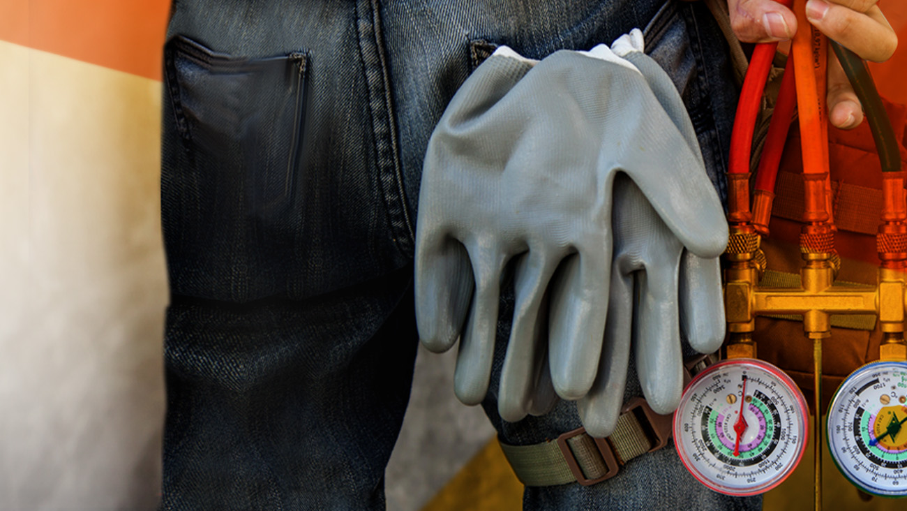 Close-up of a worker wearing jeans, holding thick rubber gloves and a manifold gauge set with red hoses. The background is a blurred mix of orange and gray tones, suggesting a work or industrial setting.