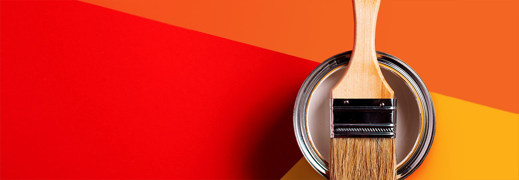 A paintbrush rests on an open can of paint against a background with large blocks of red, orange, and yellow hues. The composition creates a vibrant and colorful visual contrast.