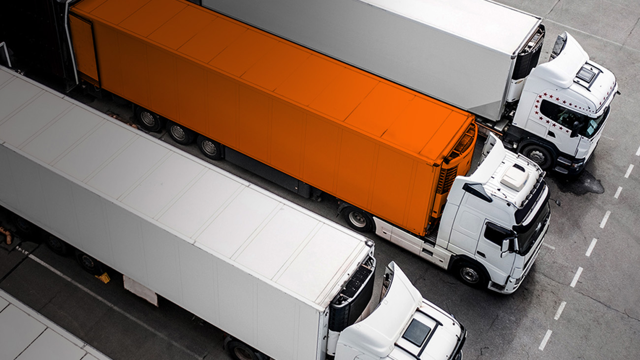 Aerial view of four parked trucks in a row, one being orange and the others white. They are aligned diagonally on a marked gray asphalt surface.