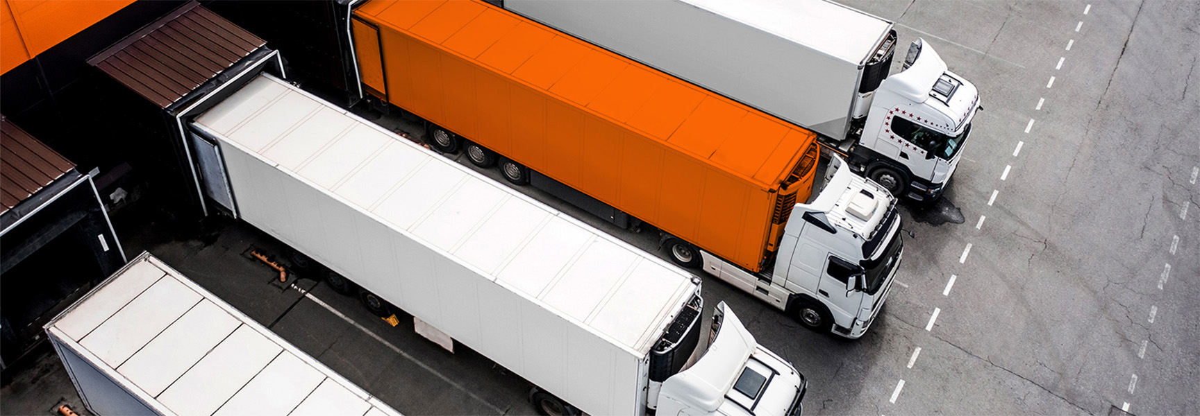 Five semi-trucks parked in a loading dock, viewed from above. Four trucks have white trailers, while one in the center has an orange trailer. The trucks are aligned in a row next to the building.