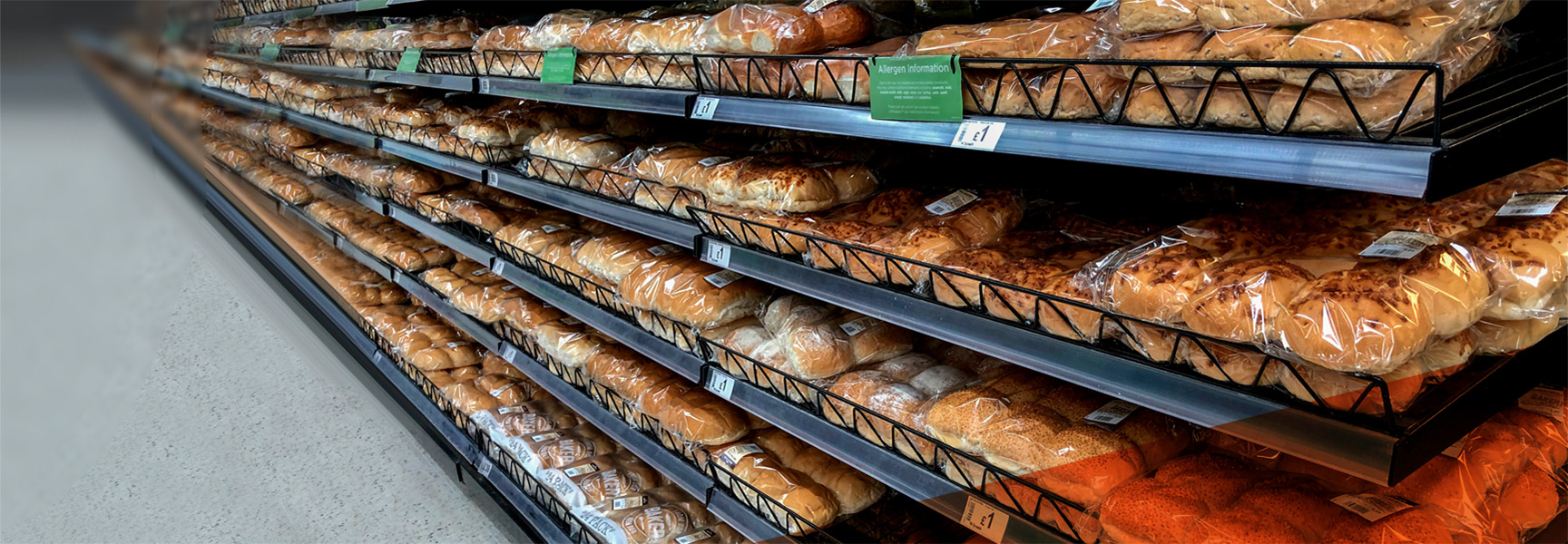 Shelves filled with various types of packaged bread rolls in a bakery section of a grocery store. The bread is neatly arranged in metal racks, and each package has a label. The floor is a light-colored tile.