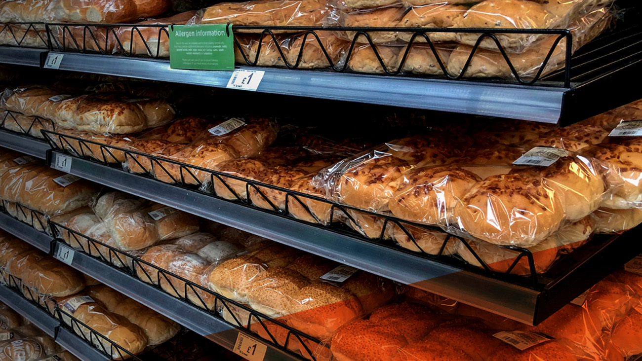 Shelves filled with various breads wrapped in plastic packaging at a bakery or supermarket. A green allergen information sign is visible above the top shelf. Each item is labeled with a price tag showing £1.