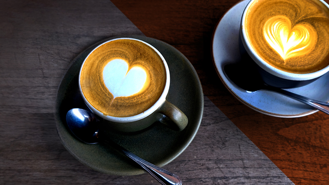 Two cups of cappuccino with heart-shaped latte art sit on saucers. One is on a wooden table with a spoon beside it. The background tables lighting contrasts between a dark wood and a lighter brown surface.