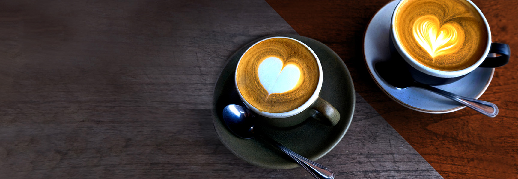 Two cappuccinos with heart-shaped latte art sit on saucers with spoons on a wooden table. The cups are placed diagonally opposite each other, one on a green saucer and one on a gray saucer. The background has a soft gradient effect.