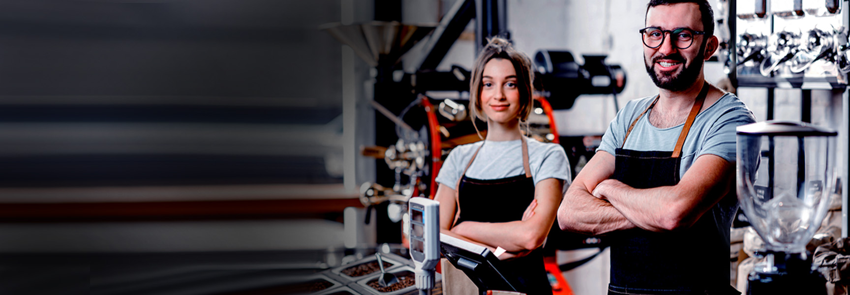 Two baristas in aprons stand smiling behind a coffee counter, surrounded by coffee-making equipment and grinders. The background is a blend of warm industrial elements and coffee machines. The scene conveys a welcoming coffee shop atmosphere.