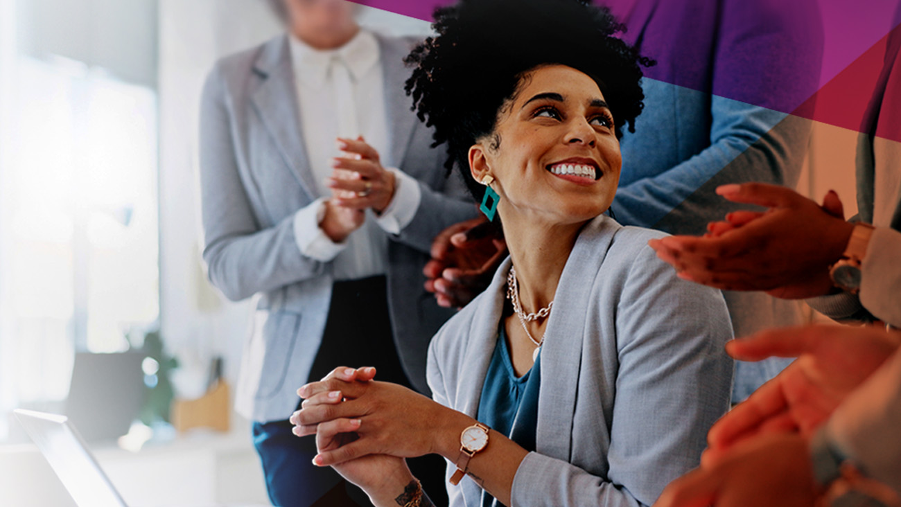 A smiling woman with curly hair sits at a table, looking up. She is surrounded by several people clapping, all wearing business attire. A laptop is visible in front of her. The atmosphere appears celebratory and supportive.