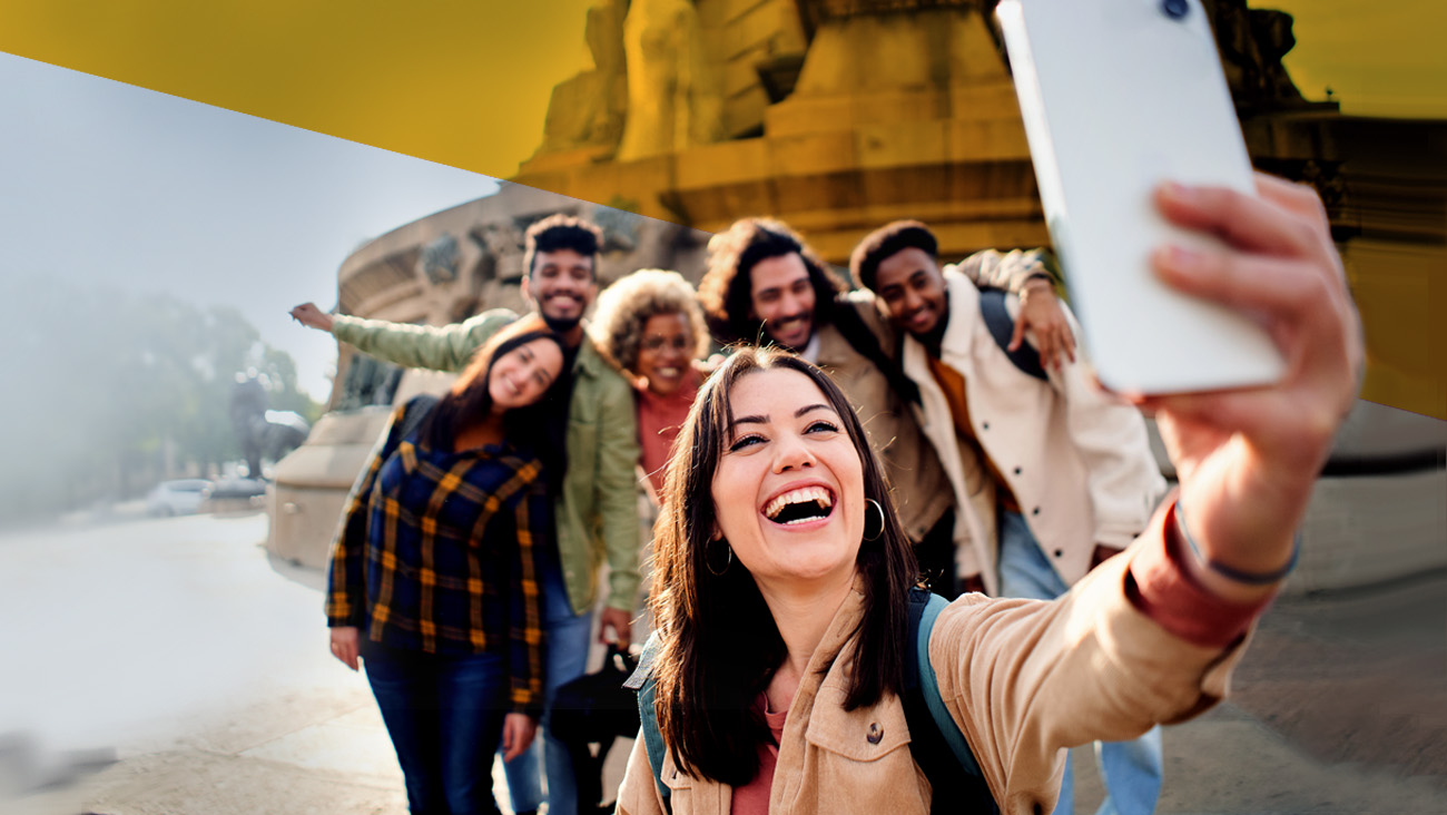 A smiling group of six friends poses for a selfie in an urban setting. The woman in front holds the phone, capturing everyone’s cheerful expressions. They are wearing casual clothing, and a historic monument is visible in the background.