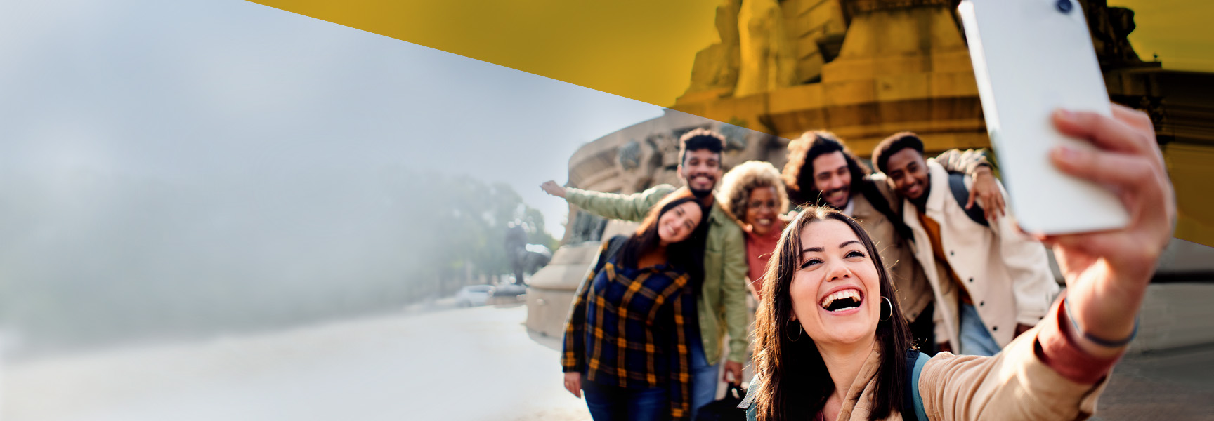 A group of six people taking a selfie outdoors, all smiling and posing together. The background shows a large architectural structure, with a painterly effect and blurred areas. The people appear to be having a joyful moment.