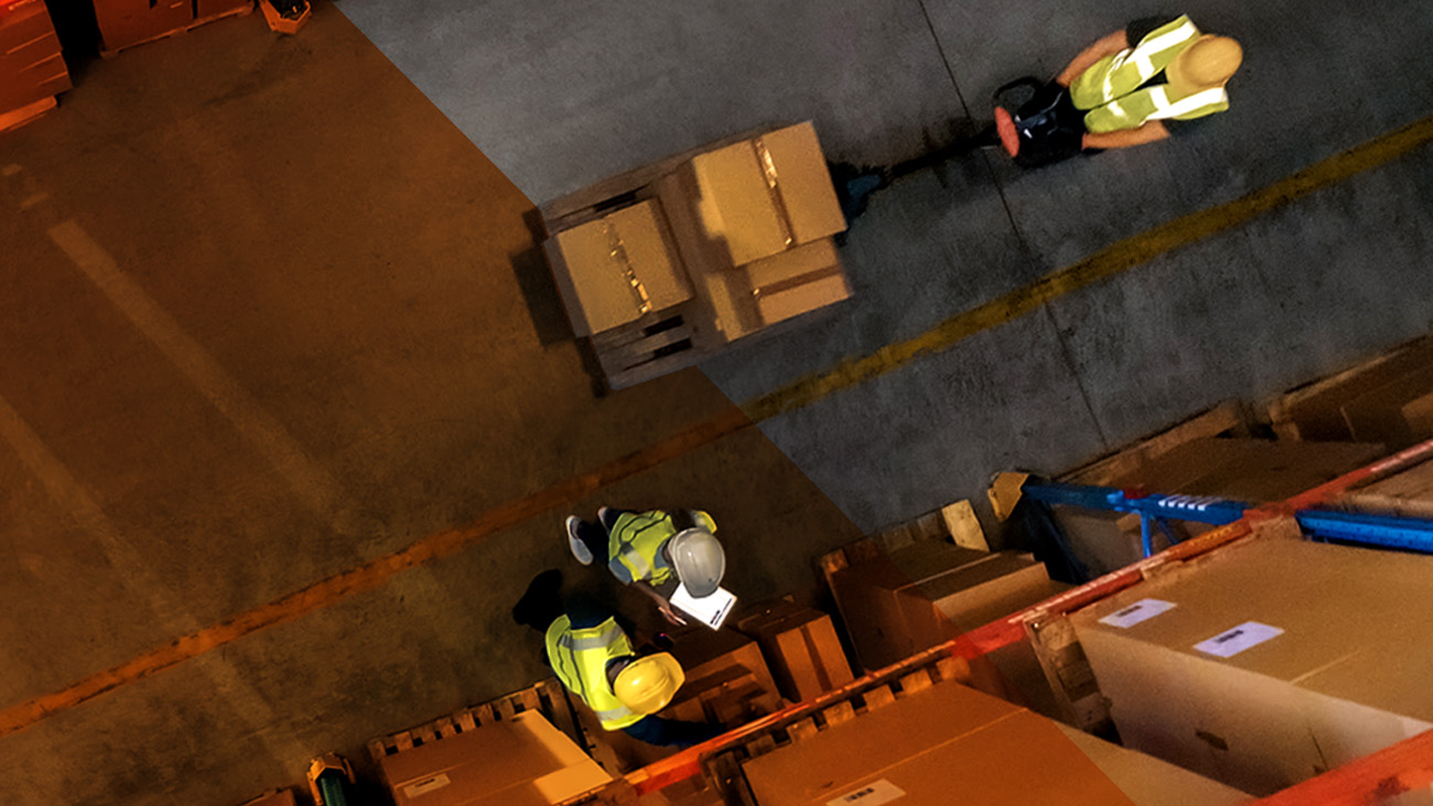 Aerial view of a warehouse with workers wearing safety vests and helmets. One worker pushes a pallet jack with boxes, while two others examine inventory. Boxes and crates are stacked along the aisles on a concrete floor.