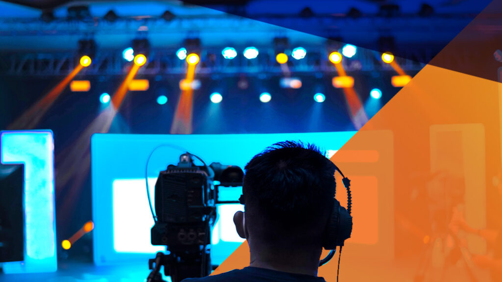 A person wearing headphones operates a camera in a studio with bright stage lights in the background. The scene is predominantly blue with a translucent orange overlay on the right side. The background shows out-of-focus illuminated screens.