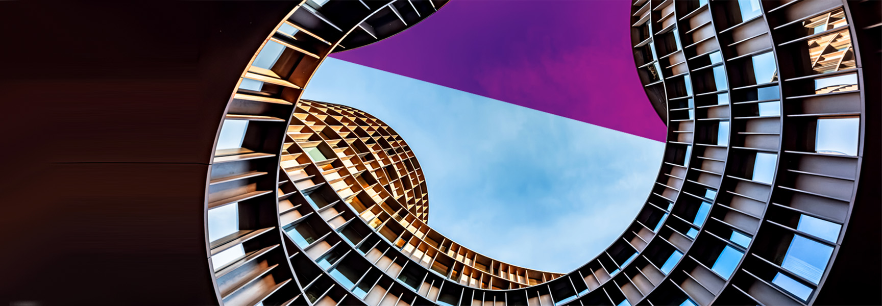 View looking up at the interior of a modern, curving building with a circular opening. The sky is visible with a mix of light blue and a purple shade. The structure features reflective windows and a futuristic design.