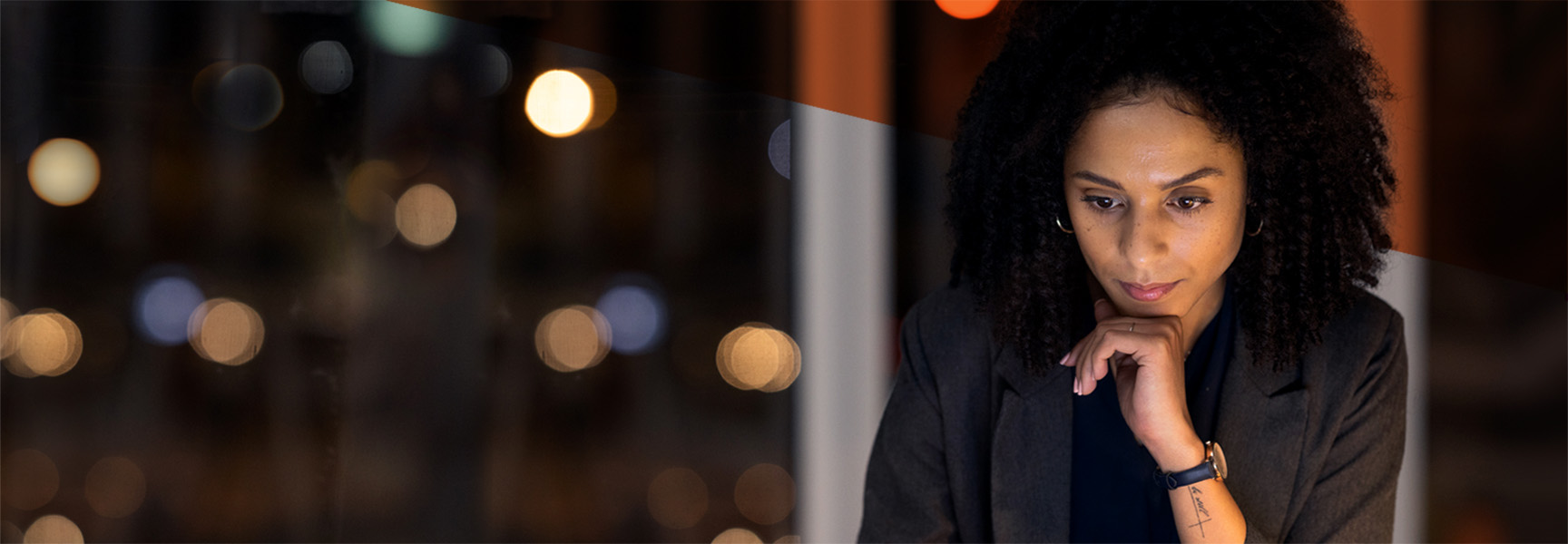 A person with curly hair and wearing a blazer is seated indoors, focusing intently on a task. The background is softly blurred, with circular lights creating a bokeh effect.