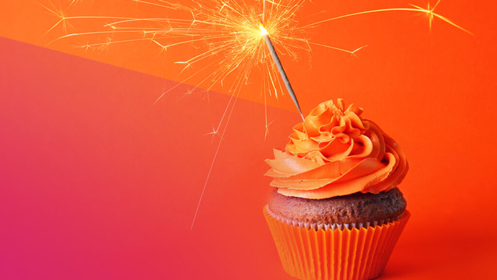 A cupcake with orange frosting is topped with a lit sparkler. The background is a gradient of pink to orange, complementing the cupcakes vibrant icing.