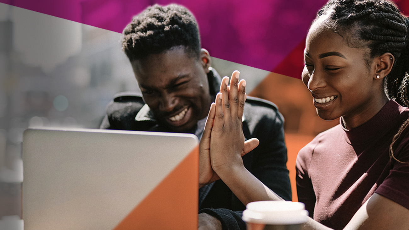 Two people sit at a table with a laptop, smiling and high-fiving. One person holds a coffee cup. The background features abstract pink and orange shapes.