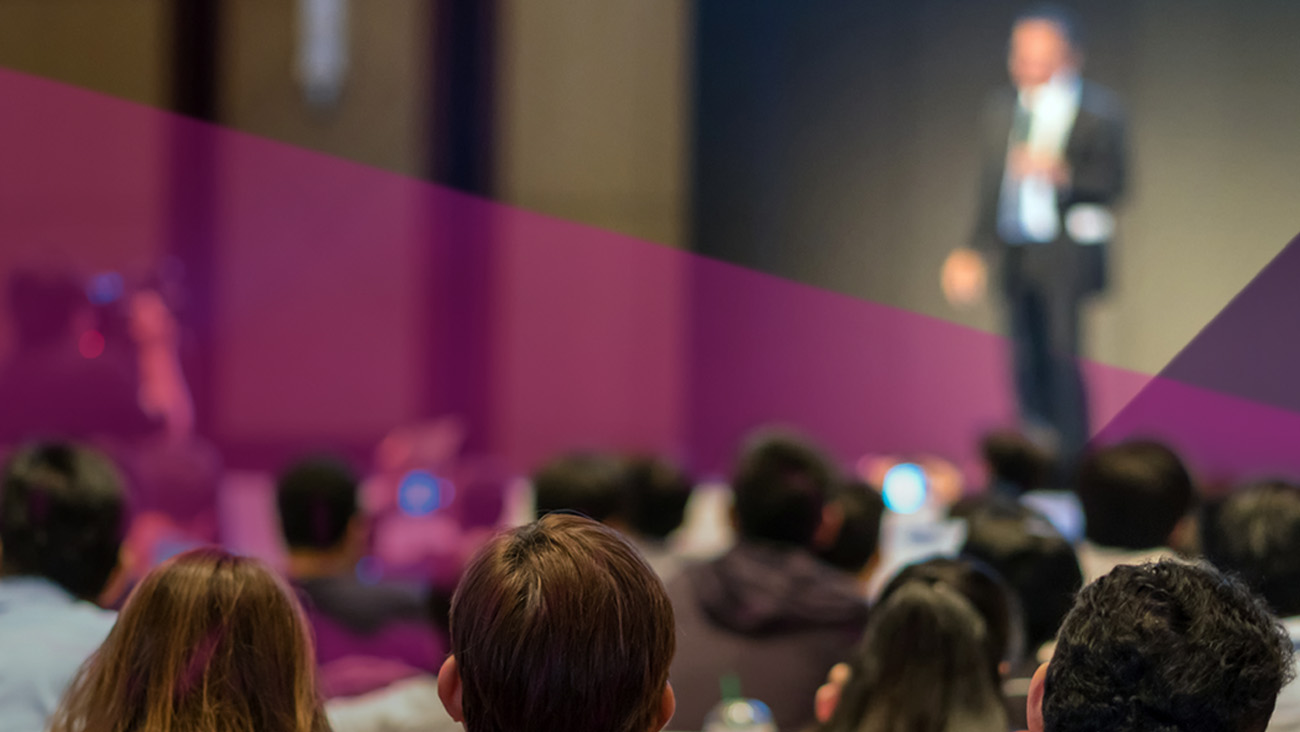 Blurry image of a speaker in a suit onstage, holding a microphone, addressing an audience. The room is dimly lit, with attendees mostly out of focus, suggesting a conference or presentation setting.