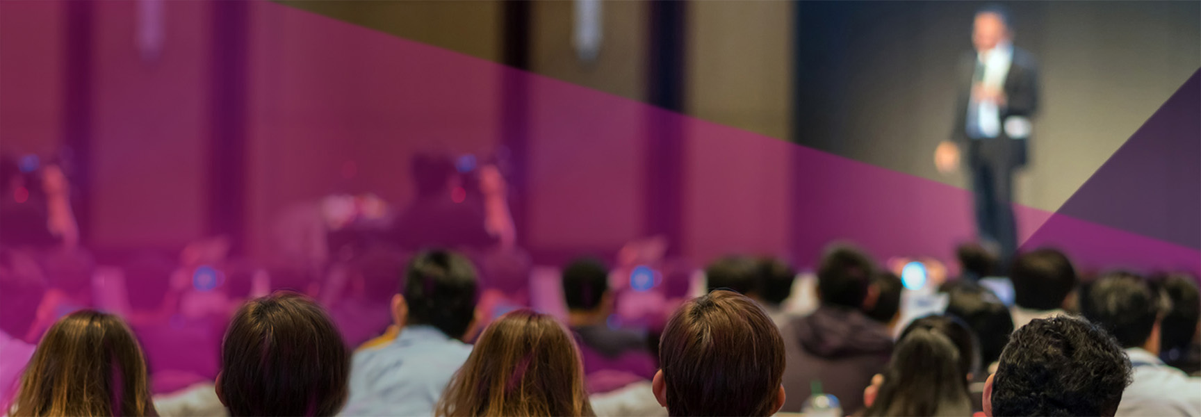 Audience seated and listening to a speaker on stage, who is blurred and speaking into a microphone. The image has a purple overlay and a professional setting with people facing forward.