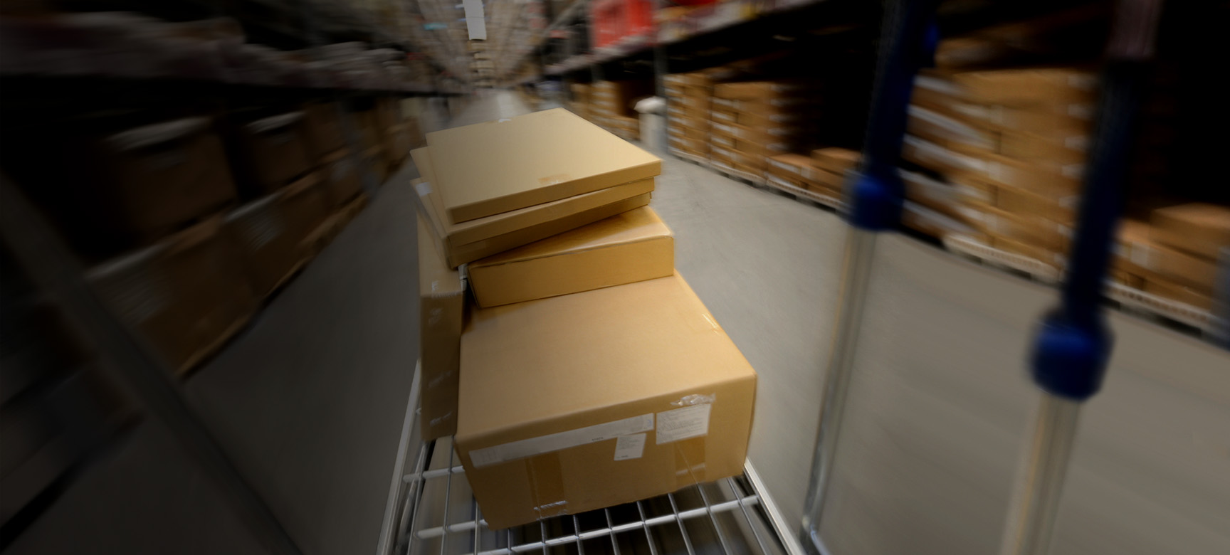 A blurry image of a warehouse aisle with stacked cardboard boxes on a moving cart. The background is filled with shelves containing various packages, suggesting fast movement.