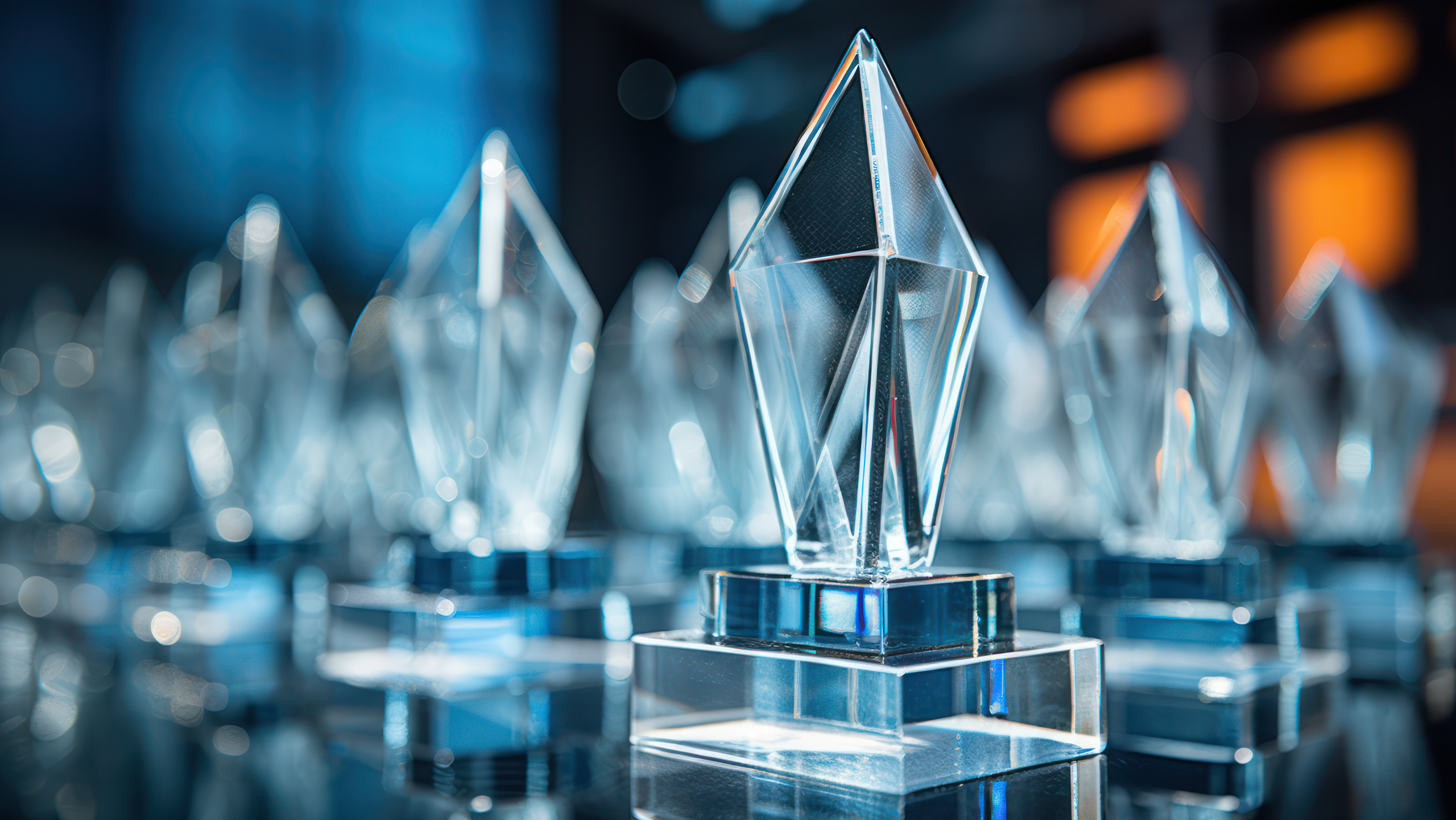 A row of transparent crystal awards with pointed tops on reflective bases. The background is softly blurred, showing hints of blue and orange lights, creating an elegant and celebratory atmosphere.