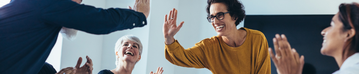 A diverse group of four people in a bright room share a joyful moment. One person in a yellow sweater gives a high-five to someone out of frame, embodying the spirit of employee appreciation quotes as the others smile and applaud, creating a warm, celebratory atmosphere.