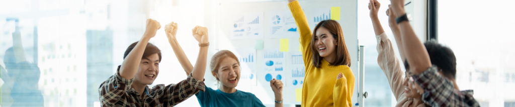 A diverse group in casual attire cheers and raises their fists joyfully in an office setting, embodying the spirit of employee appreciation. Charts and graphs on a board indicate a successful presentation, while bright natural light fills the spacious, modern room.