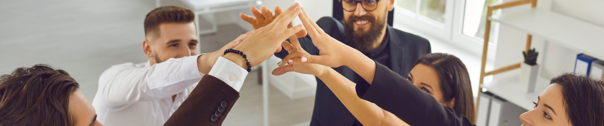 A group of people stands in a circle, reaching their hands in to form a team huddle. They are smiling, emanating positivity and collaboration under bright office lights, embodying the spirit of employee appreciation quotes that celebrate teamwork and camaraderie.