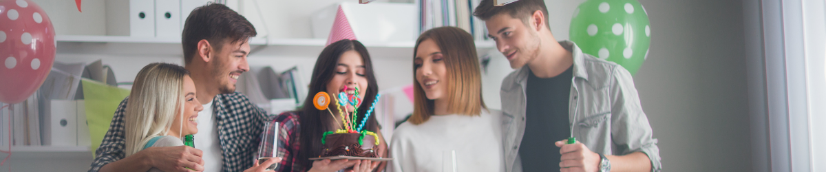 A group of five people celebrates a birthday in a room adorned with colorful balloons and banners, embodying the spirit of employee appreciation. They gather around a chocolate cake with candles; one wears a pink party hat. Everyone looks cheerful and festive.