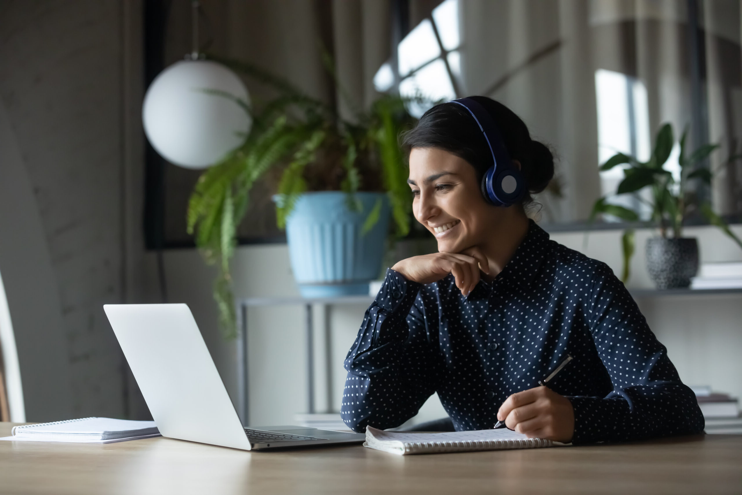 A woman with dark hair in a bun and a polka-dot shirt, headphones snugly on, smiles at her laptop as she types out employee appreciation quotes. Shes seated at a table with a notepad and pen nearby, framed by a cozy ambiance of a plant and lamp in the background.