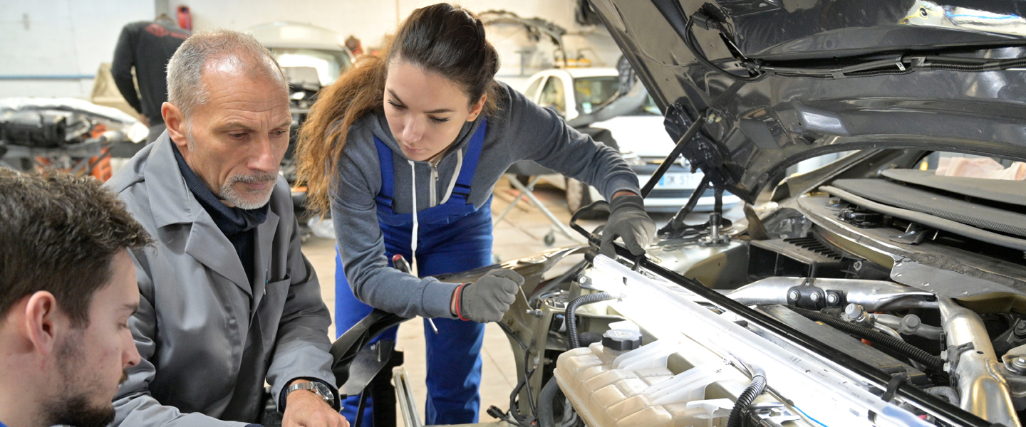 A group of three people, two men and a woman, are engaged in automotive learning as they work on a car engine in the garage. The woman, wearing work gloves and coveralls, seems to receive instruction from the older man. Various tools and cars fill the background.