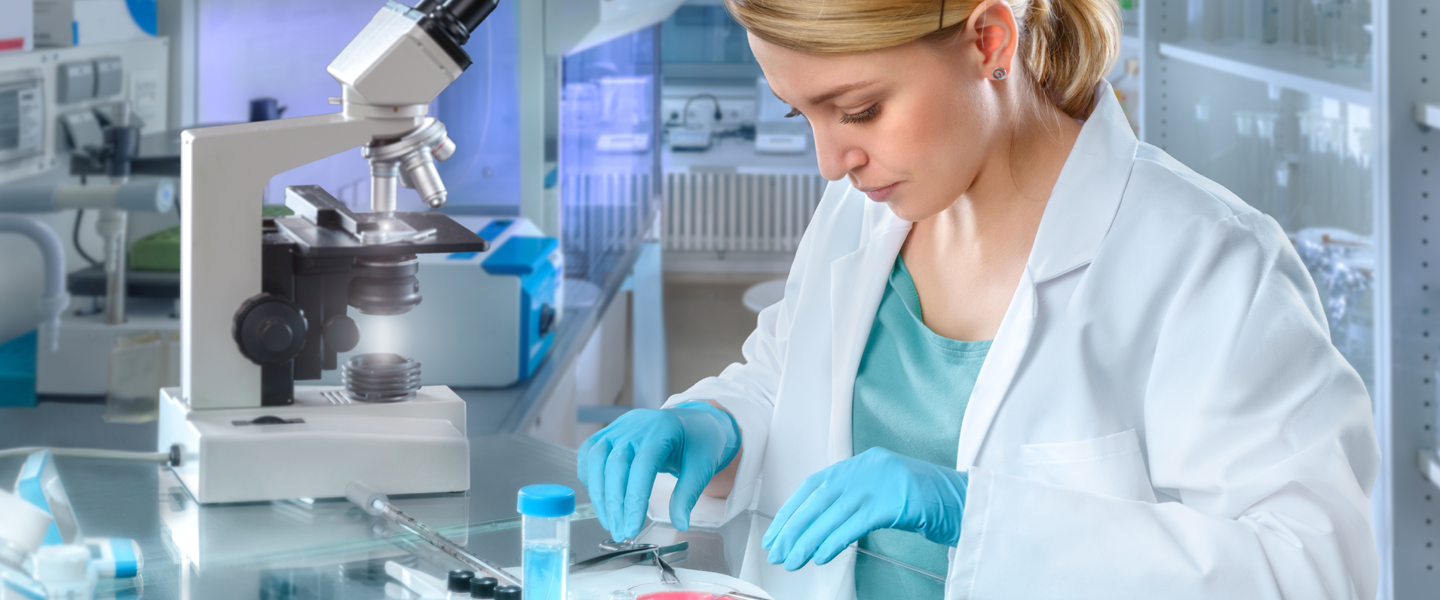 A scientist in a lab coat and gloves examines a petri dish at a laboratory workstation, keeping her focus sharp amidst the bustling environment. With a microscope and various lab equipment surrounding her, she calmly navigates the labs vibrant scene where sometimes, threes a crowd.