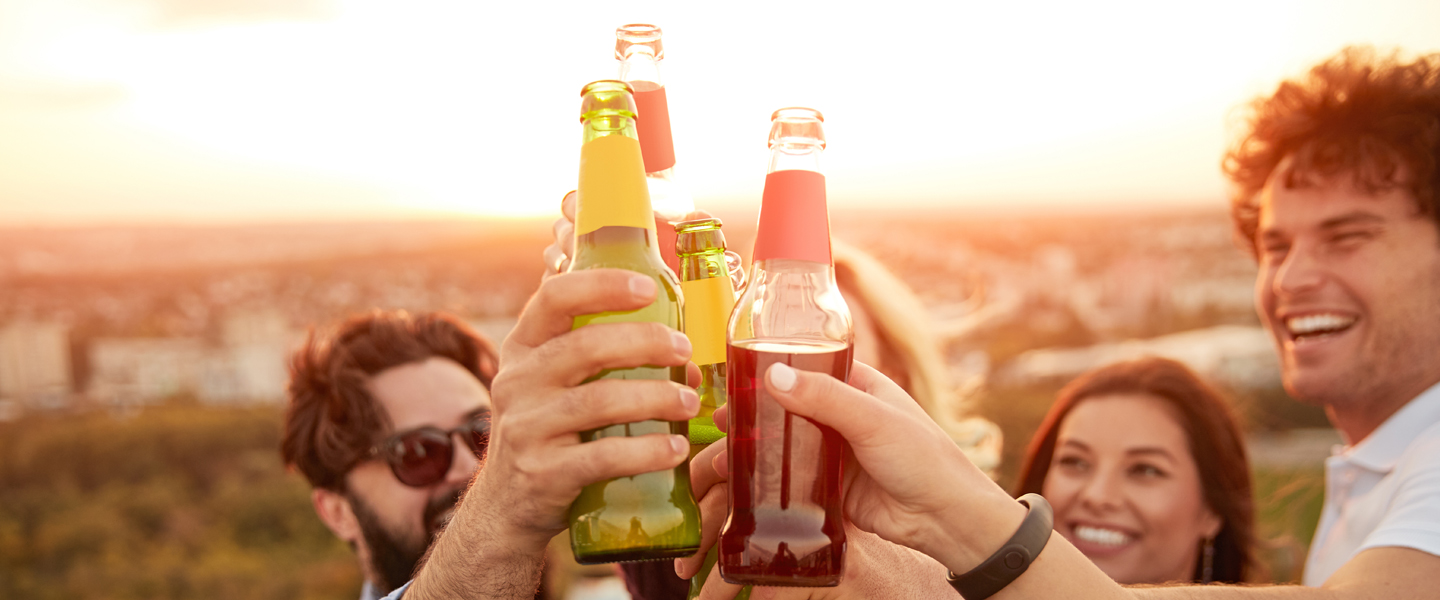 A group of people raise bottles in celebration, with a sunset casting a warm glow over their gathering. Smiling faces suggest a joyful atmosphere as they embrace the spirit of travel and adventure, with distant cityscape views enhancing this outdoor moment.