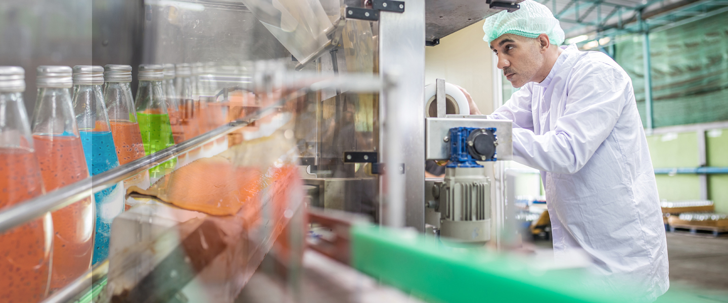 A worker in a white coat and hairnet operates machinery in a beverage factory, highlighting a multi-channel production approach. Various colored liquid-filled bottles line the conveyor belt, emphasizing the industrial focus on efficient and diverse beverage output.