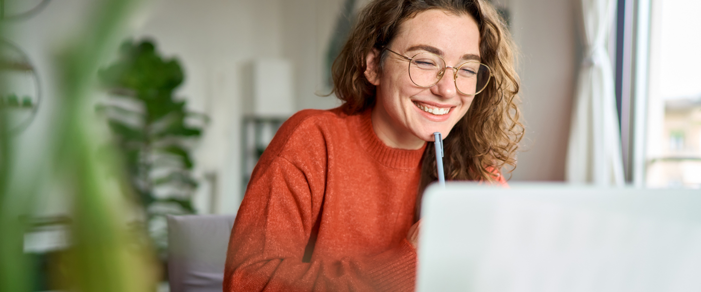 A person with curly hair and glasses, wearing an orange sweater, smiles while looking at a laptop, perhaps engaged in an employee learning program. They hold a pen near their face in the bright room filled with natural light, and a green plant adds a touch of nature in the background.