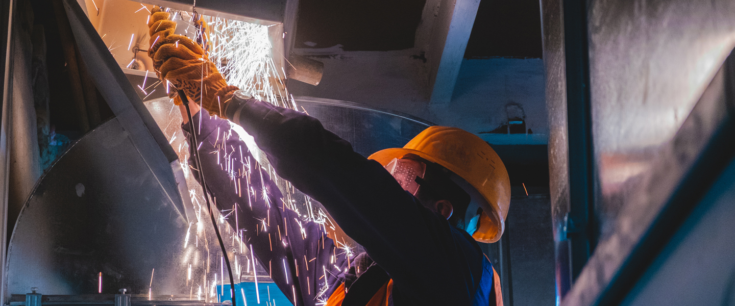 A worker in a hard hat and protective gear is welding metal, causing a shower of bright sparks. The industrial scene, with HVAC systems and machinery surrounding the worker, comes alive as the lighting highlights the sparks against the dim backdrop.