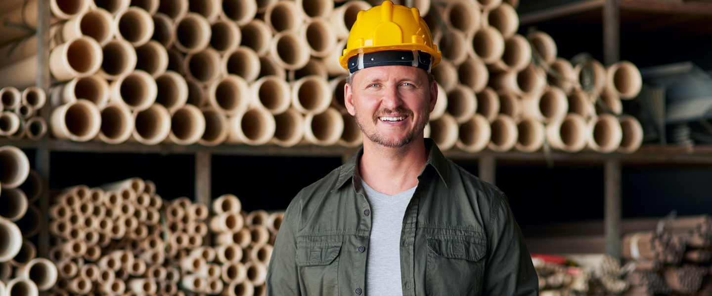 A smiling contractor, known for his loyalty, wears a yellow hard hat and a green jacket as he stands before shelves stocked with stacked PVC pipes in the warehouse.