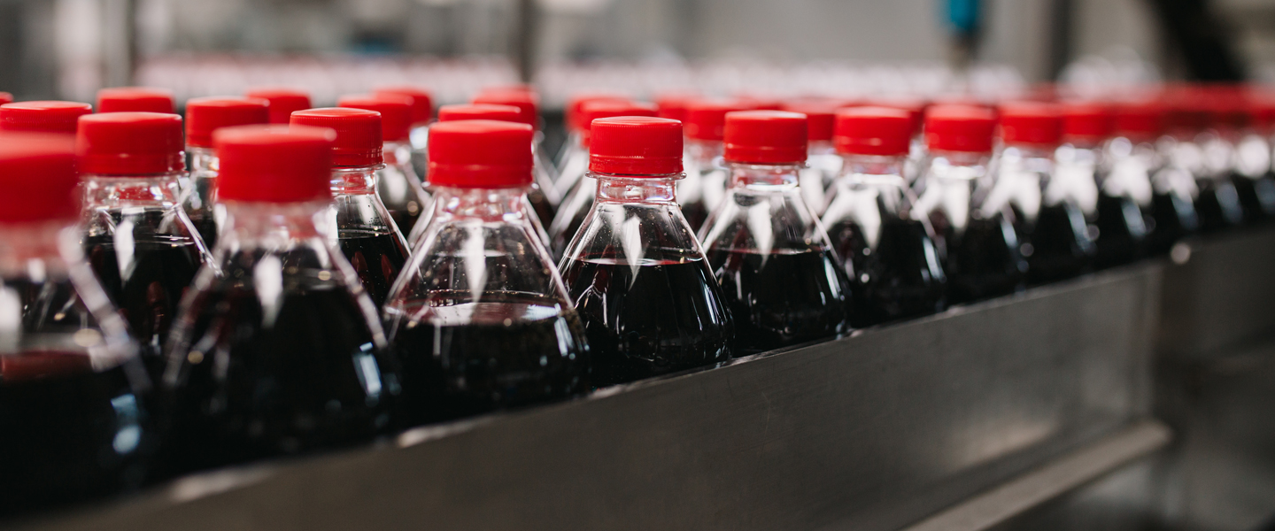 Rows of plastic bottles with red caps, filled with a dark liquid likely to be soda, are lined up on a conveyor belt in a bottling facility, leading the way in efficiency and precision.