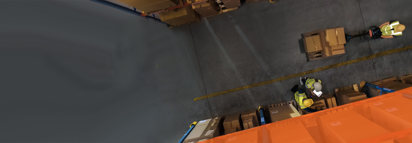 Aerial view of a bustling warehouse scene with two workers in yellow safety vests, enhancing channel engagement as they efficiently handle brown cardboard boxes. Boxes are neatly stacked on shelves and the floor, with shadows and a distinct yellow safety line guiding the workspace flow.