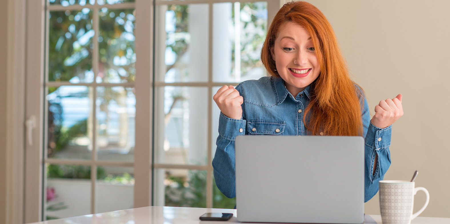 A red-haired woman in a denim shirt sits at a table, smiling and clenching her fists in excitement while looking at a laptop. Amidst sunny windows, shes thrilled by the potential of using gamification to drive behaviors. A smartphone and coffee mug rest on the table.