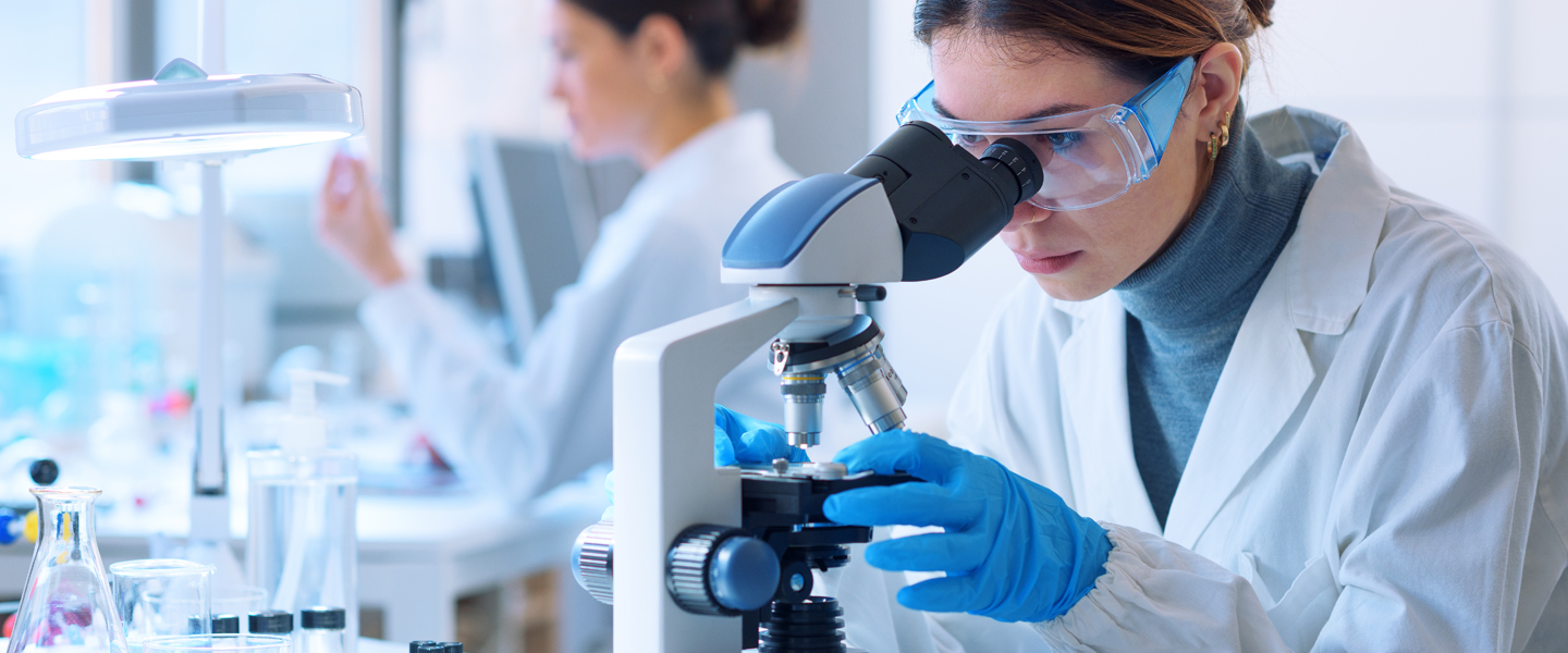 A scientist wearing a lab coat, safety goggles, and blue gloves looks into a microscope in a laboratory. Another scientist is in the background, also working. The environment is bright and equipped with lab equipment.