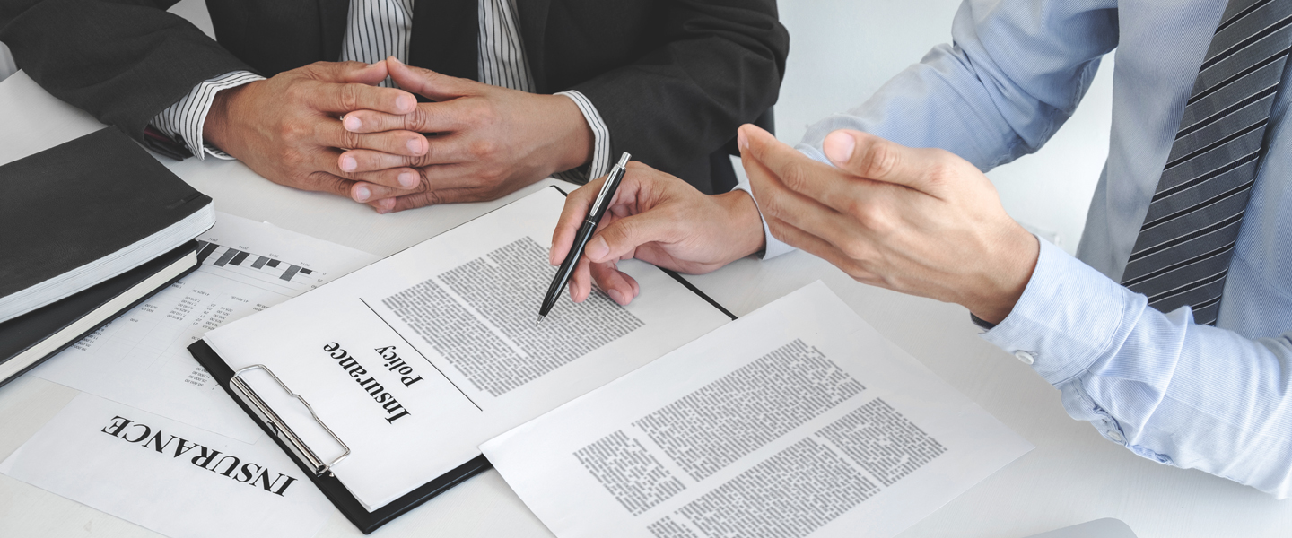 Two individuals in business attire are seated at a table, reviewing an insurance policy document with their insurance agent. One person is holding a pen over the document while the other gestures with a smartphone in hand. Nearby are various papers and a laptop.