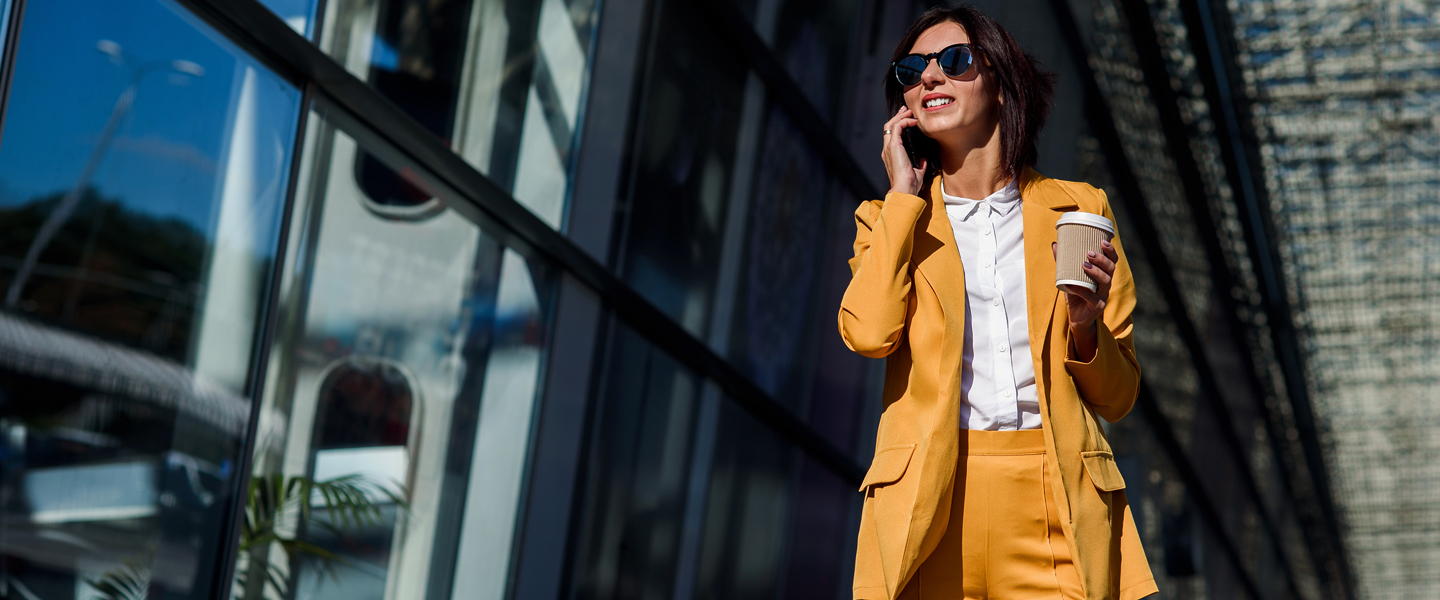 A woman in a yellow suit and sunglasses walks outside a glass building, phone in one hand and coffee cup in the other. Her targeted approach to morning routines shines through as the sun casts reflections on the glass around her.