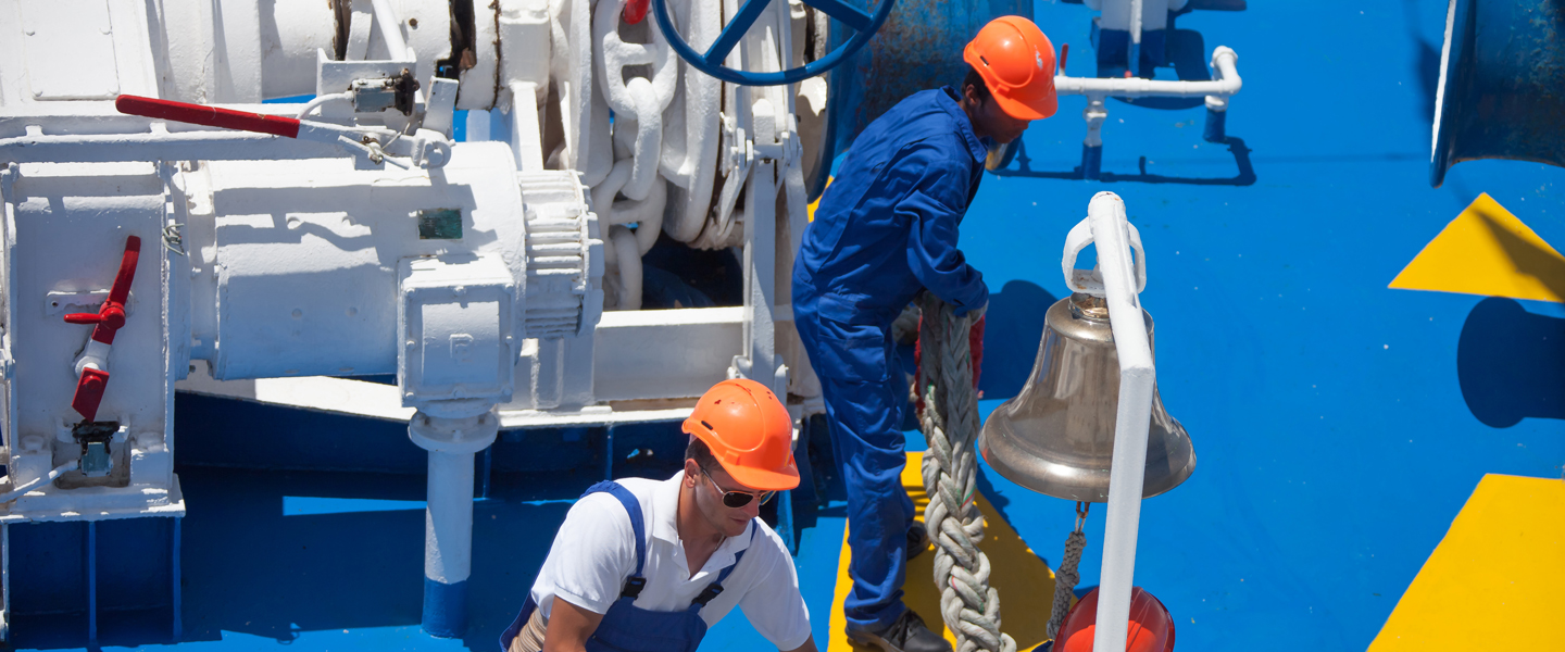 Two workers in orange helmets and blue overalls are handling a thick rope on a blue deck next to a large ship bell. Machinery and parts of the boat are visible in the background.