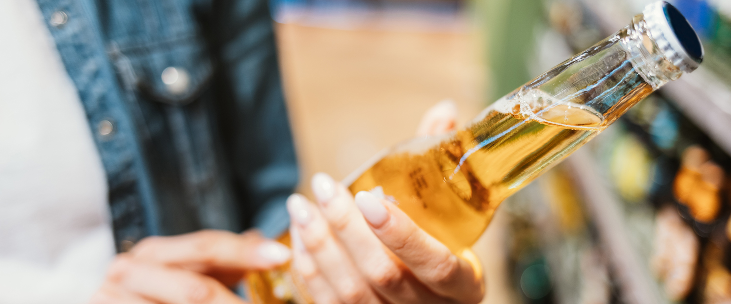 A person, tapping into success, holds a glass bottle filled with a light golden beverage, possibly beer, in a store. The hand deftly adjusts the bottle cap as shelves brimming with various products frame the scene in the background.