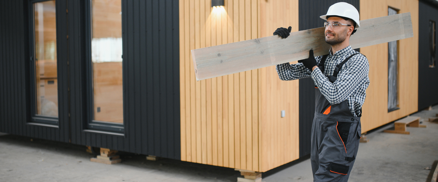 A construction worker in a white hard hat and overalls carries a wooden plank on his shoulder. He is walking past a modern building with black and wooden paneling.