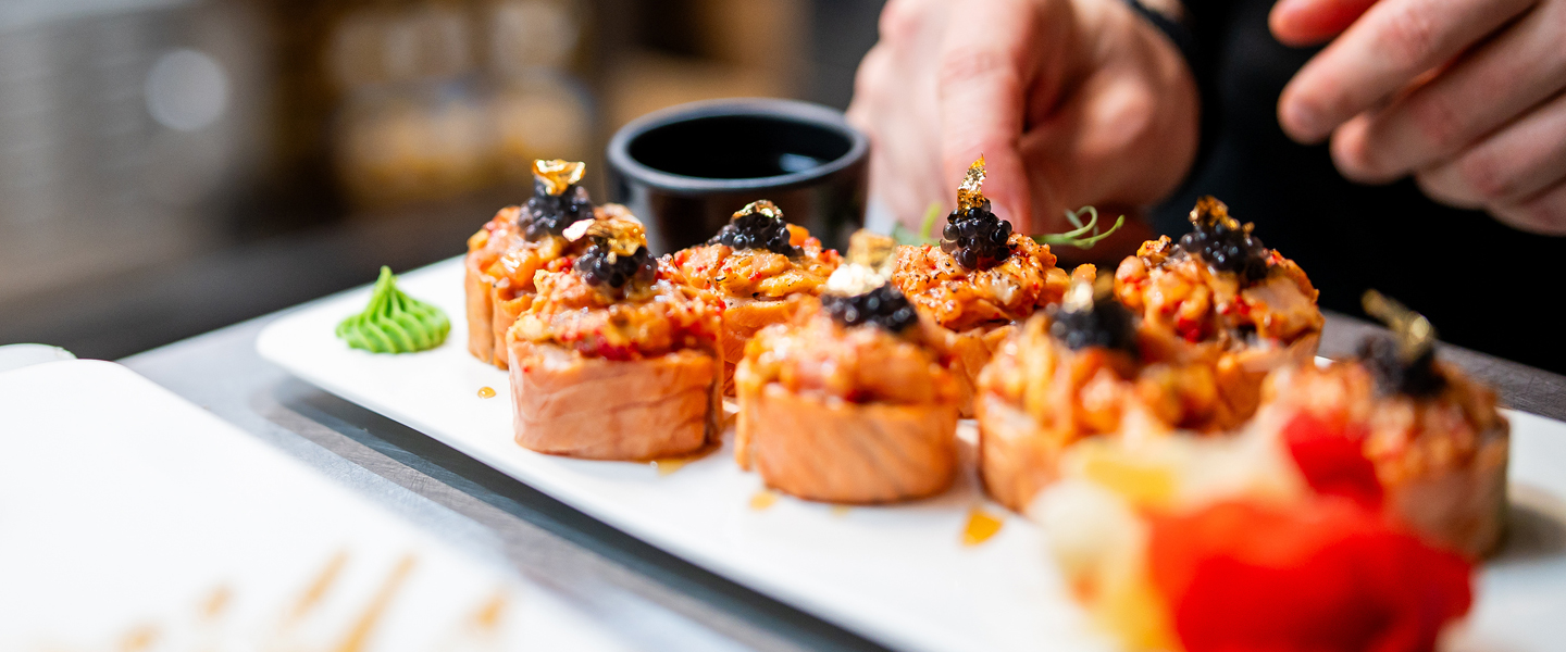 Close-up of gourmet sushi rolls topped with gold flakes and caviar on a white plate, perfectly arranged by a chefs hands. Ideal for group travel dining experiences, a small black bowl and green garnish add to the exquisite presentation in the background.